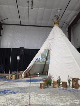 A 30-foot tall tipi decorated with fireweed and wood logs around. Inside, two projectors mapped along the tipi's canvas displaying vibrant, immersive visuals.