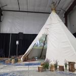 A 30-foot tall tipi decorated with fireweed and wood logs around. Inside, two projectors mapped along the tipi's canvas displaying vibrant, immersive visuals.